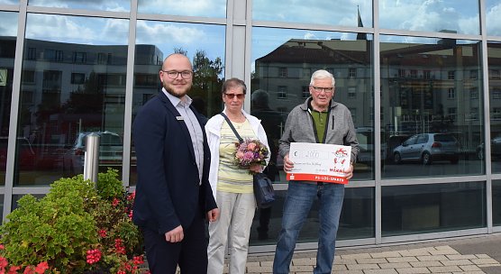 Kundenberater Jonas Echtermeier &uuml;berreicht den gl&uuml;cklichen Gewinnern einen symbolischen Scheck und Blumen. (Foto: KSK)